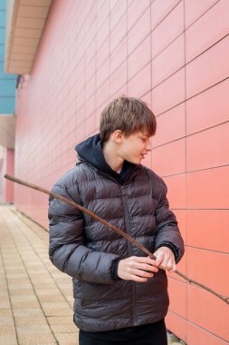 Koyu renk kabarık ceketli genç çocuk, duvarın yanında uzun bir dal tutuyor, modern şehir ortamını seyreden bir gencin samimi yaşam tarzı portresi.
