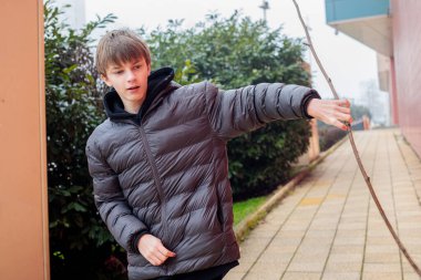 Koyu renk kabarık ceketli gülümseyen genç bir çocuk elinde uzun bir dalla şehir ortamında sopaya bakan neşeli ergen portresi.