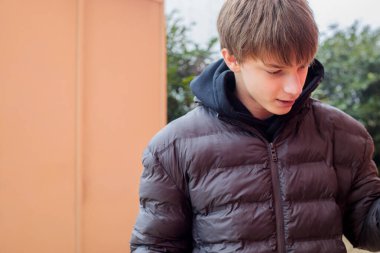 Koyu renk kabarık ceketli ve kapüşonlu bir çocuk açık havaya bakıyor. Güpegündüz, şehir arka planına sahip düşünceli bir gencin doğal samimi portresi.