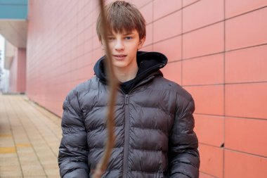 Siyah ceketli, kendine güveni tam genç bir çocuk modern kırmızı bir duvarın yanında duruyor, şehir ortamında ciddi ve şık görünüyor.