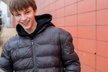 Siyah ceketli, gülümseyen genç bir çocuk modern bir duvarın yanında duruyor, mutlu ve huzurlu görünüyor.