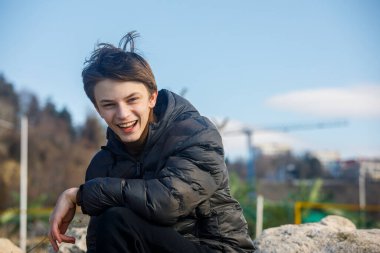 Açık havada koyu renk ceketli gülümseyen bir gencin fotoğrafı, yumuşak bulanık arka planda kameraya bakıyor. Doğal ışık, gündelik yaşam tarzı konsepti
