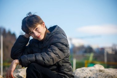 Açık havada koyu renk ceketli gülümseyen bir gencin fotoğrafı, yumuşak bulanık arka planda kameraya bakıyor. Doğal ışık, gündelik yaşam tarzı konsepti