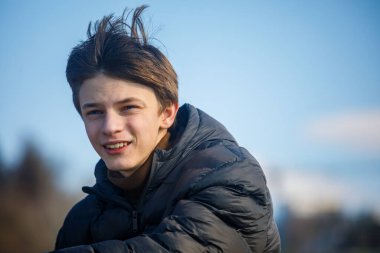 Açık havada koyu renk ceketli gülümseyen bir gencin fotoğrafı, yumuşak bulanık arka planda kameraya bakıyor. Doğal ışık, gündelik yaşam tarzı konsepti