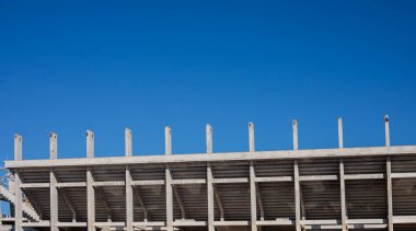 Açık mavi gökyüzü altındaki beton stadyum yapısı, dikey kolonları ve geometrik çizgileri olan boş tribünler, gündüz kentsel mimari arka planı