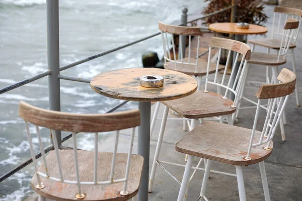 Yıpranmış deniz kenarı kafe terası boş ahşap sandalyeler ve su kenarında küçük yuvarlak masalar dalgalı sahil geçidi ve kasvetli atmosfer.