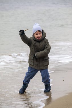 Deniz dalgaları yakınındaki sahilde oynayan küçük çocuk elinde sopayla serin sahil yürüyüşünde açık hava aktivitelerinin keyfini çıkarıyor.