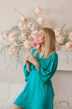 Yeşil elbiseli zarif bir kadın, çiçek açan güllerin yanında pembe çiçekleri tutuyor, bahar havasını ve rafine pastel iç kısımda kadın gününü kutluyor.