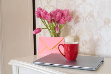 Kırmızı kahve kupası laptopta ofis masasında pembe laleler ve zarfla, uzaktan kumandalı çiçeklerle, ilkbahar ya da yaz işgünü sabah kahve molası