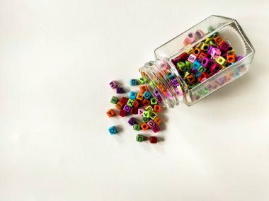 A glass jar lies on its side, spilling colorful alphabet beads onto a light surface. 