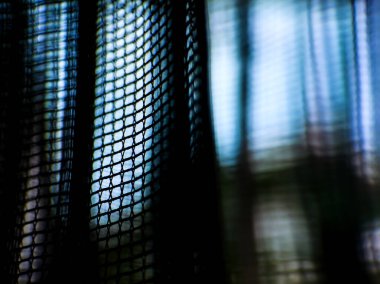 Aydınlık bir pencereye karşı koyu renkli bir ağ perdesinin yakın görüntüsü, koyu, ağlı kumaş ve parlak mavi ışık arasında bir kontrastlık yaratır.. 