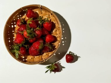 Yuvarlak, dokuma ahşap bir sepet içinde canlı kırmızı derili ve yeşil yapraklı olgun çilekler var.. 