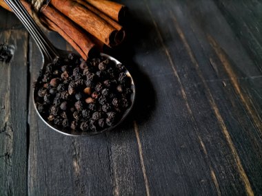 Bir metal kaşık, koyu ahşap bir zemin üzerinde sayısız siyah karabiber taşır ve kırsal bir zemin oluşturur.. 