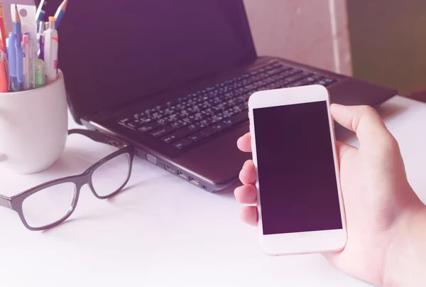 Closeup hand hold smartphone and laptop on desk background community device in office or home, soft focus. — Stock Photo, Image