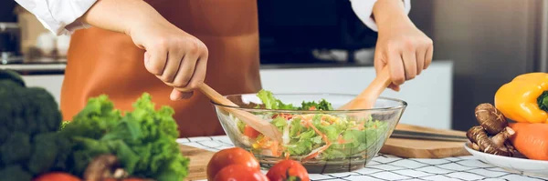 Asian woman is mixing the ingredients in a salad bowl at the kitchen cooking table.