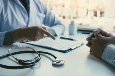 Doktor, elinde kalem tutarak hastanın geçmişini not defterine yazıyor ve hastayla ilaç ve tedavi hakkında konuşuyor..