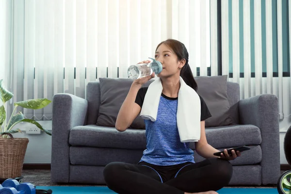 Asian woman sportswoman drinking from water bottle in her room after yoga exercise at home.
