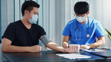 Asyalı doktor, yıllık kontrolü sırasında bir hastanın tansiyon monitörünü kullanıyor ve kan basıncını açıklıyor..
