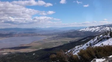 Bulutlar ve gölgeler bir çöl vadisinde karlı tepelerin üzerinden geçiyor evler ve ağaçlarla çevrili bir göl.