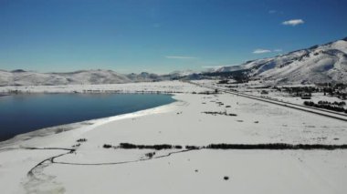 Drone pan left over winter lake shores with snowy mountain reflections