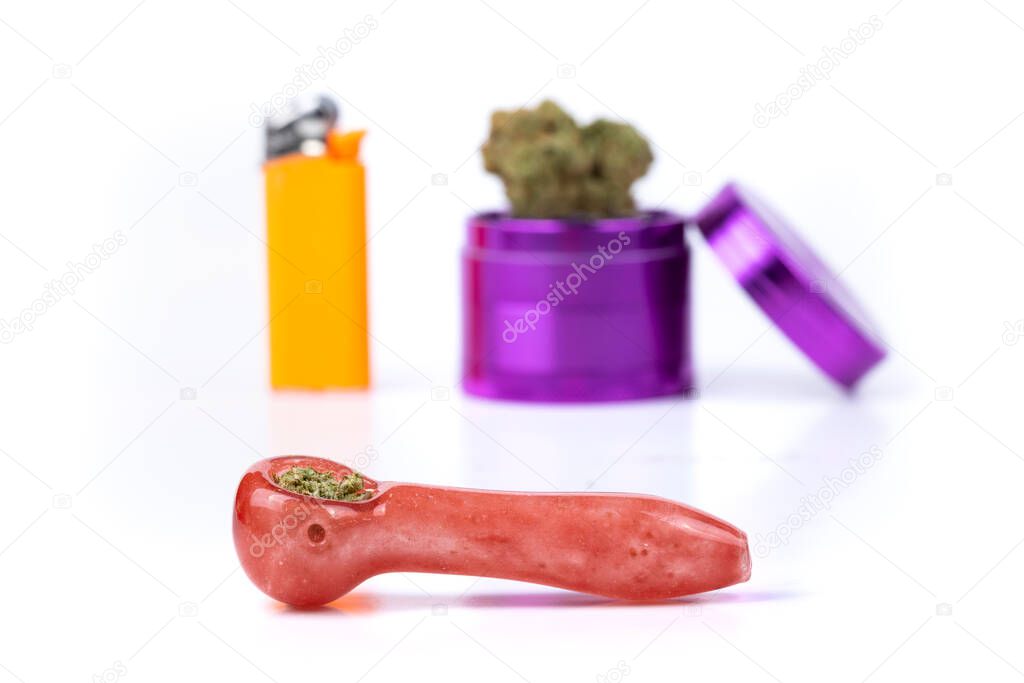 In focus salmon colored glass pipe filled with weed out of focus background with grinder, bud and lighter