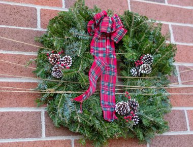 Süslü Noel çelengi, kırmızı ekose bir fiyonk, çam kozalakları ve tuğla bir duvarda asılı böğürtlen. Doğal yemyeşil dallardan yapılan bayram süsü..