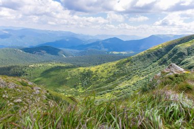 Kayalık yeşil yamaçları, sık ormanları ve bulutlu bir yaz gökyüzünün altındaki uzak mavi tepeleri olan panoramik bir dağ manzarası. Yürüyüş parkurundan manzara manzarası.