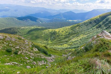 Bereketli yeşil yamaçları, kayalık arazisi ve açık bulutlu gökyüzünün altındaki uzak mavi tepeleri olan manzaralı bir dağ vadisi. Yazın yürüyüş parkurundan güzel doğa manzarası