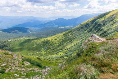 A scenic mountain valley with lush green slopes, rocky terrain and distant blue peaks under a bright cloudy sky. Beautiful nature view from a hiking trail in summer