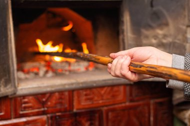 Close-up of human hands using a wooden tool to tend fire inside a traditional brick oven. Artisan bread baking, rustic cooking, home oven and traditional food preparation concept.