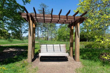 Güneşli bir günde yeşil bir parkta pergola 'nın altında sallanan ahşap salıncak bankı. Mavi gökyüzü ile huzurlu bir açık hava rahatlama arkaplanı.