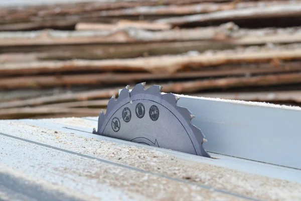 Profesyonel bir testereye yerleştirilmiş keskin dairesel testere bıçağının yatay yakın çekimi. Bıçakta güvenlik simgeleri var ve yüzey yeni talaşlarla kaplı. Bulanık arka planda, tahta desteler görünür, temsil eder