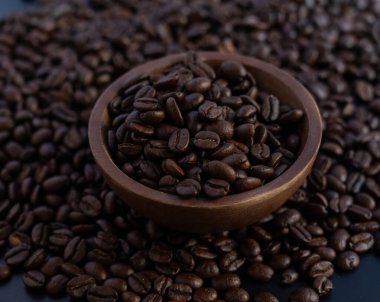 El yapımı ahşap bir kâsenin içinde gölgeler ve dokuları vurgulayan koyu bir arka planda fasulye yığınıyla çevrili zengin kavrulmuş kahve çekirdeklerinin sinematik sade bir fotoğrafı..