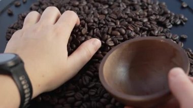 Bir kişinin ellerinin büyük bir kazıktan taze kavrulmuş kahve çekirdeklerini kepçeleyip küçük el yapımı ahşap bir kaseye koyduğunu gösteren 4K sinema videosu. Harekete ve dokuya odaklı sessiz görüntüler.