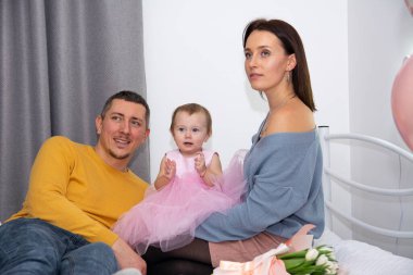 Genç bir aile: anne, baba ve küçük sevimli bir kız çocuğu pembe elbiseli beyaz bir yatakta, ve onların yanında çiçekler var..