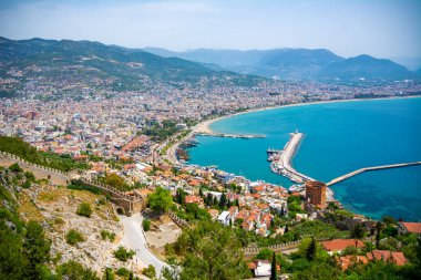Yukarıdan Alanya şehrinin güzel manzarası. Yeşil ağaçlara gömülü kadim bir savunma duvarı ve bir tarafında mavi deniz, diğer tarafında dağlar olan bir tatil köyü..