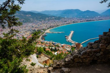 Yukarıdan Alanya şehrinin güzel manzarası. Yeşil ağaçlara gömülü kadim bir savunma duvarı ve bir tarafında mavi deniz, diğer tarafında dağlar olan bir tatil köyü..