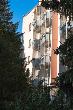 Kırmızı balkonları ve yanında köknar ağaçları olan çok katlı bir bina. Balkonları olan bir Alman apartmanı. Bazı balkonlarda çiçekler ve şemsiyeler vardır..