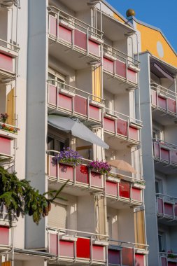 Kırmızı balkonları ve yanında köknar ağaçları olan çok katlı bir bina. Balkonları olan bir Alman apartmanı. Bazı balkonlarda çiçekler ve şemsiyeler vardır..