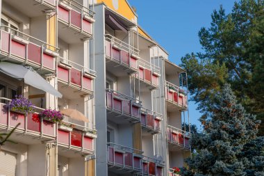 Kırmızı balkonları ve yanında köknar ağaçları olan çok katlı bir bina. Balkonları olan bir Alman apartmanı. Bazı balkonlarda çiçekler ve şemsiyeler vardır..