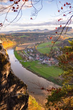 Sakson İsviçre, Bastei Doğa Koruma Alanı. Panoramik nehir manzarası, güzel bir Alman kasabası, güneş ışığıyla çevrili canlı bir sonbahar ormanı, ufukta dağlar ve bulutlu bir gökyüzü..