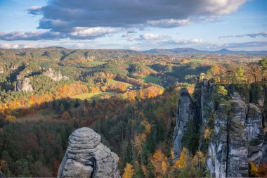 Sakson İsviçre, Bastei Doğa Koruma Alanı. Güneş ışığında canlı bir sonbahar ormanı, ufukta dağlar ve bulutlu bir gökyüzü ile çevrili güzel bir Alman köyünün panoramik manzarası..