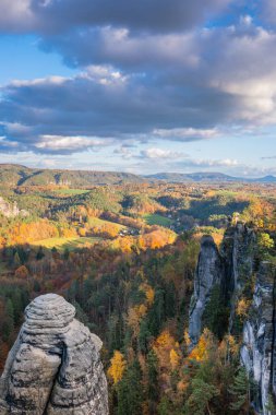 Sakson İsviçre, Bastei Doğa Koruma Alanı. Güneş ışığında canlı bir sonbahar ormanı, ufukta dağlar ve bulutlu bir gökyüzü ile çevrili güzel bir Alman köyünün panoramik manzarası..