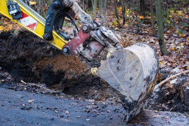 Çamurla kaplı küçük bir kazıcı kovası asfalt bir yolda duruyor. Parkta kazı çalışmasında bir mola.
