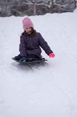 Karlı bir tepeden kayan küçük bir kız parlak bir kar kaydırağıyla. Soğuk bir günde, bir orman parkında aktif kış oyunlarının keyfini çıkaran mutlu bir çocuk..