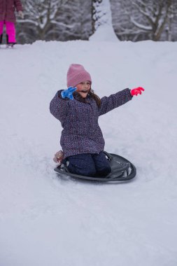 Karlı bir tepeden kayan küçük bir kız parlak bir kar kaydırağıyla. Soğuk bir günde, bir orman parkında aktif kış oyunlarının keyfini çıkaran mutlu bir çocuk..