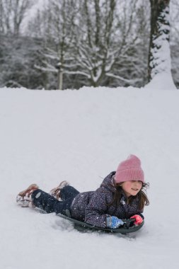 Karlı bir tepeden kayan küçük bir kız parlak bir kar kaydırağıyla. Soğuk bir günde, bir orman parkında aktif kış oyunlarının keyfini çıkaran mutlu bir çocuk..