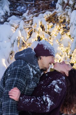 Karla kaplı güzel bir ormanda kucaklaşan ve el ele tutuşan genç bir çift. Kar yağışı sırasında romantik kış yürüyüşü. Kışlık ceketli kadın ve erkek, karla dolu uzun çam ağaçlarının arasında huzurlu bir anın tadını çıkarıyorlar.