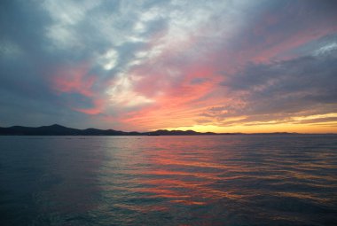 Ufukta Ugljan Adası 'nın karanlık silueti olan Adriyatik Denizi' nin üzerinde mavi bir saat gün batımı. Turuncu, pembe ve mor tonlarla boyanmış dramatik bulutlar sakin suyu yansıtıyor ve huzurlu bir deniz manzarası yaratıyor. Sahne kıyı duvar kâğıdı.
