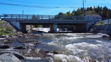 Kırsal beton bir köprünün altından geçerek merkez sütuna ulaşan çağlayan bir nehrin üzerinden alçak seviye uçuş. Riviere-Blanche, Saint-Ulric-de-Matane, Quebec, Kanada, 2025.
